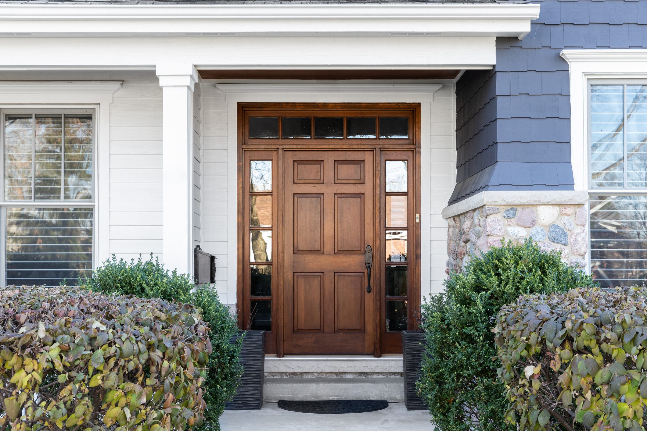 front porch of a modern home featuring a durable wooden door with glass panels on each side, framed by white siding and stone accents, surrounded by neatly trimmed bushes, showcasing energy-efficient and secure door installation by Midwest Best Exteriors & More