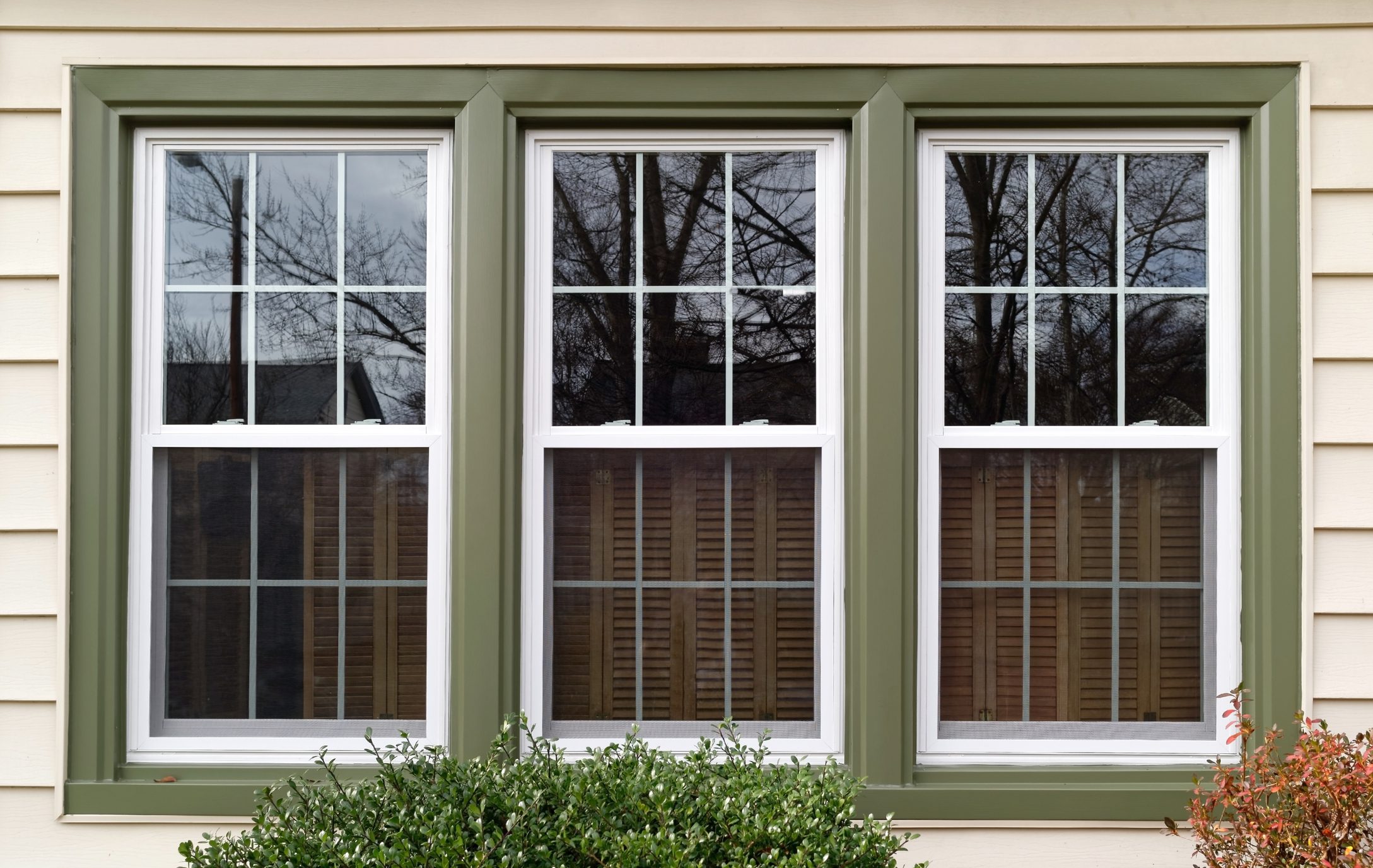 Three energy-efficient white-framed double-hung windows with green trim installed on a beige house exterior, reflecting trees and sky, showcasing durable, stylish window solutions for improved home insulation and curb appeal.